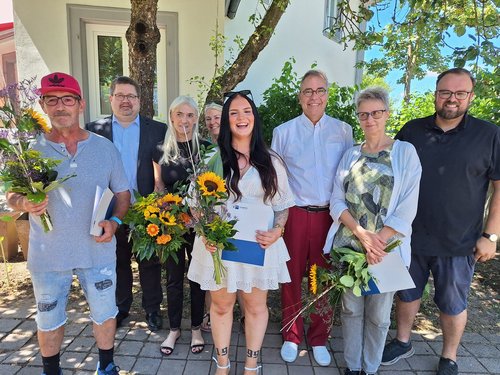 Ein Gruppenfoto von acht Personen im Freien. Sie halten Blumensträuße mit Sonnenblumen und Ordner. Eine junge Frau im weißen Kleid steht lächelnd im Zentrum. Die Szene deutet auf eine Verleihung oder feierliche Anerkennung hin.