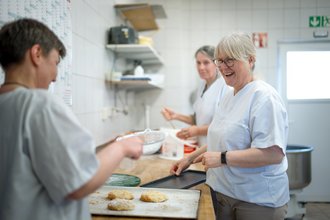 Drei Frauen in weißen Arbeitskleidern backen gemeinsam in einer Küche. Eine lächelnde Frau mit Brille zeigt auf ein Backblech mit Gebäck, während eine Kollegin im Vordergrund arbeitet. Im Hintergrund rührt eine weitere Frau in einer Schüssel. Die Szene wirkt freundlich und arbeitsam.