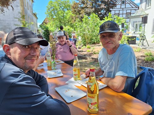 Zwei Männer sitzen an einem Holztisch im Freien und lächeln in die Kamera. Auf dem Tisch stehen Flaschen Apfelschorle und leere Teller. Im Hintergrund sind weitere Personen, Bäume und Gebäude bei sonnigem Wetter zu sehen.