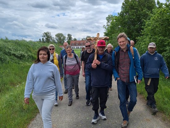 Eine Gruppe von Menschen wandert auf einem Kiesweg. Ein Mann in der Mitte trägt ein großes Holzkreuz auf der Schulter. Sie bewegen sich durch eine ländliche Umgebung mit grünen Feldern und Bäumen unter einem bewölkten Himmel.
