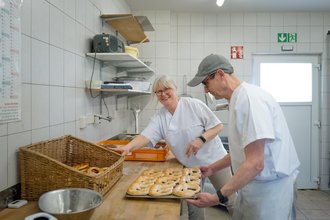 Zwei Personen in weißer Arbeitskleidung stehen in einer Küche. Eine lächelnde Frau mit Brille greift in einen Korb, während ein Mann mit Kappe ein Blech mit frischen Bagels hält.