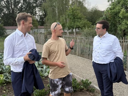 Drei Männer unterhalten sich auf einem Kiesweg in einem Garten. Der Mann in der Mitte in Freizeitkleidung gestikuliert, während die beiden Männer in Hemden lächelnd zuhören. Im Hintergrund sind Bäume und ein Holzzaun zu sehen.