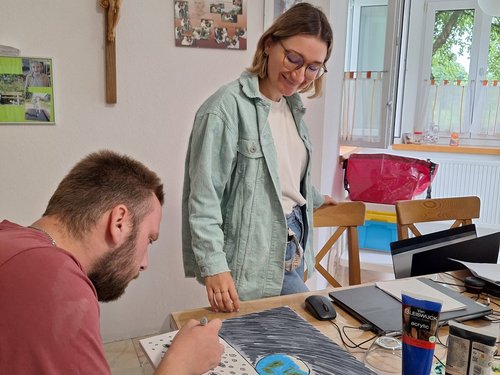 Ein bärtiger Mann malt an einem Tisch ein Bild mit einem blauen Planeten und einer Mondlandschaft. Eine Frau mit Brille steht lächelnd daneben und beobachtet ihn. Auf dem Tisch liegen Acrylfarben.