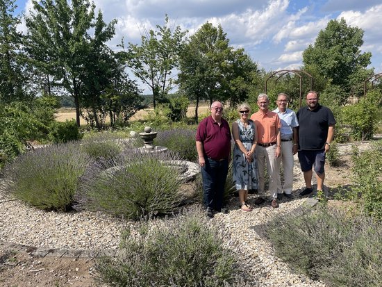 Fünf Personen stehen auf einem Kiesweg in einem Garten, umgeben von Lavendelsträuchern und Bäumen. Im Hintergrund ist ein Brunnen und ein blauer Himmel zu sehen. Die Gruppe posiert gemeinsam für ein Foto.