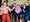 Eine Gruppe fröhlicher Menschen tanzt in einem Saal. Im Vordergrund tanzen eine Frau in roter Hose, ein Mann im rosa Hemd und ein Mann im blauen Pullover lachend nebeneinander.