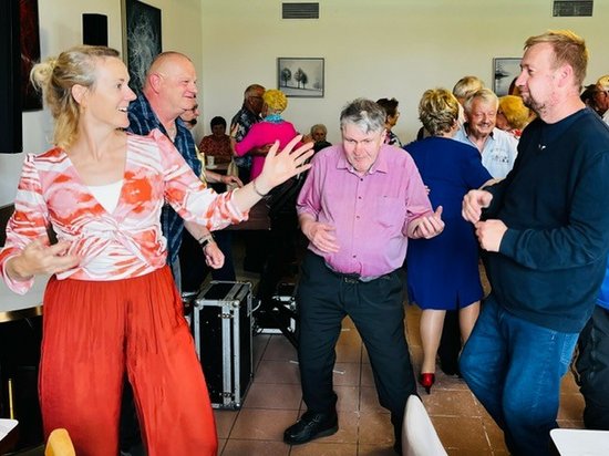Eine Gruppe fröhlicher Menschen tanzt in einem Saal. Im Vordergrund tanzen eine Frau in roter Hose, ein Mann im rosa Hemd und ein Mann im blauen Pullover lachend nebeneinander.