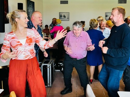 Eine Gruppe fröhlicher Menschen tanzt in einem Saal. Im Vordergrund tanzen eine Frau in roter Hose, ein Mann im rosa Hemd und ein Mann im blauen Pullover lachend nebeneinander.