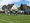 Eine Gruppe von Menschen, viele in hellblauen Shirts, ruht sich auf einem grünen Rasensportplatz aus. Einige sitzen auf dem Gras, andere stehen. Im Hintergrund sind Wohnhäuser, geparkte Autos, Fahrräder und ein Kirchturm unter einem blauen Himmel zu sehen.