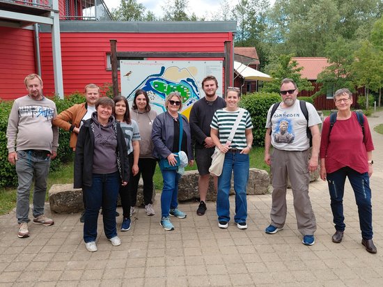 Eine Gruppe von neun Personen steht lächelnd nebeneinander im Freien vor einem roten Holzgebäude. Dahinter ist eine Informationstafel mit einer Karte angebracht. Die Personen tragen legere Freizeitkleidung.