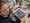 Eine Frau mit sichtbaren Tattoos sitzt an einem Tisch in einem Kunstworkshop. Vor ihr liegt ein farbenfrohes Gemälde einer Blume. Der Tisch ist mit Acrylfarben, Pinseln und Malutensilien bedeckt. Im Hintergrund arbeitet eine weitere Person an einem eigenen Bild.