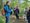 Ein Mann in einer blauen Jacke mit Rucksack steht auf einem Waldweg und gestikuliert, während er zu einer Gruppe von Menschen spricht, die ihm aufmerksam zuhören. Im Hintergrund sind hohe Bäume und grünes Gras zu sehen.