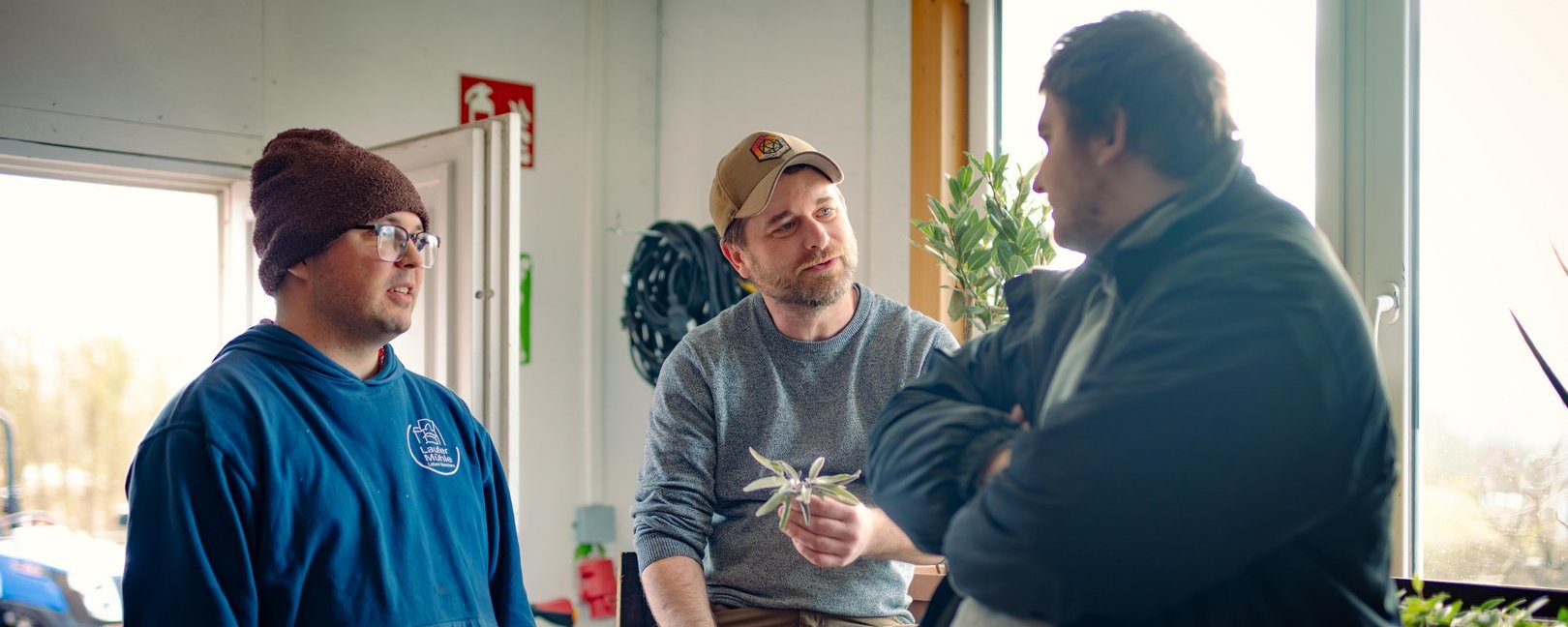 Drei Männer unterhalten sich in einer Werkstatt. Ein sitzender Mann in der Mitte hält eine kleine Pflanze und spricht mit einem Kollegen, während ein dritter Mann im blauen Hoodie links im Bild zuhört.