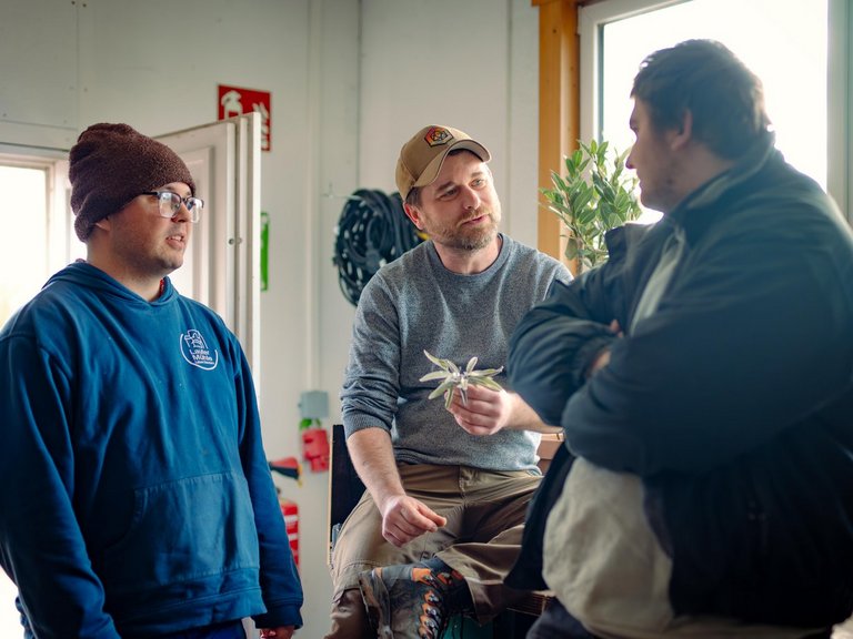 Drei Männer unterhalten sich in einer Werkstatt. Ein sitzender Mann in der Mitte hält eine kleine Pflanze und spricht mit einem Kollegen, während ein dritter Mann im blauen Hoodie links im Bild zuhört.