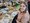 Eine lächelnde Frau mit blonden Haaren sitzt an einem Tisch voller Kunstmaterialien. Sie hält eine Collage mit Naturmotiven und dem Text „Jetzt tanze ich durchs Leben". Im Hintergrund sind Pinsel, Farben und Magazine zu sehen, was auf einen kreativen Workshop hindeutet.