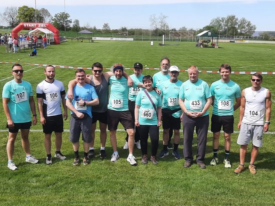 Eine Gruppe von elf Läufern posiert auf einer grünen Wiese vor einem roten Aufblasbogen mit der Aufschrift „START ZIEL“. Die meisten tragen türkisfarbene Trikots und sichtbare Startnummern.